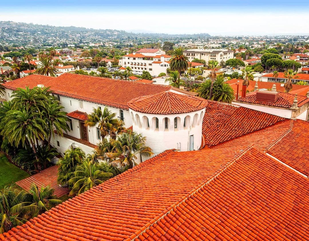 Aerial view of a red-tiled Spanish-style building surrounded by palm trees in a coastal city