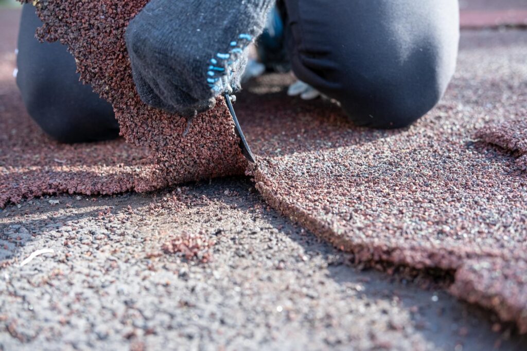 Person applying roofing material with a utility knife, focused on removing excess granules from a textured surface. The image shows close-up details of the roofing process, highlighting the texture and color of the material used.